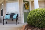 Front porch of a senior living community with a black metal bench featuring two blue patterned cushions, a green door with a wreath, a large window with closed blinds, and a neatly trimmed bush in the foreground.