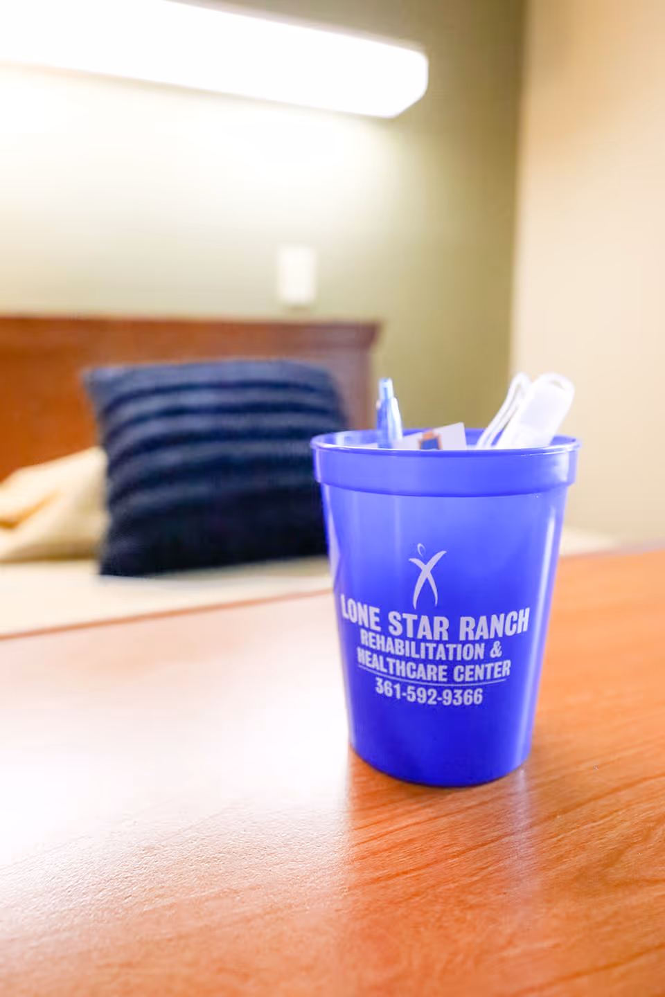 A blue plastic cup with pens and other small items inside sits on a wooden surface. The cup has the text 'Lone Star Ranch Rehabilitation & Healthcare Center 361-592-9366' printed on it. In the background, there is a blurred view of a bed with a dark blue pillow and a beige pillow against a wooden headboard.