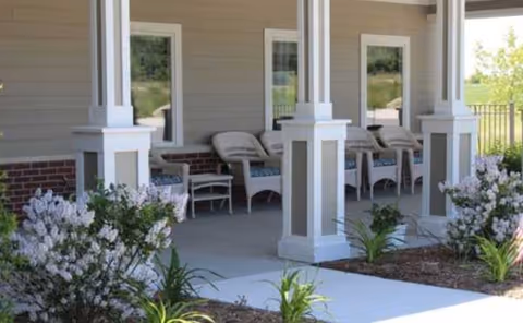 Covered porch of Harvest Home Senior Living Bancroft Haus with white columns, chairs, and shrubs.