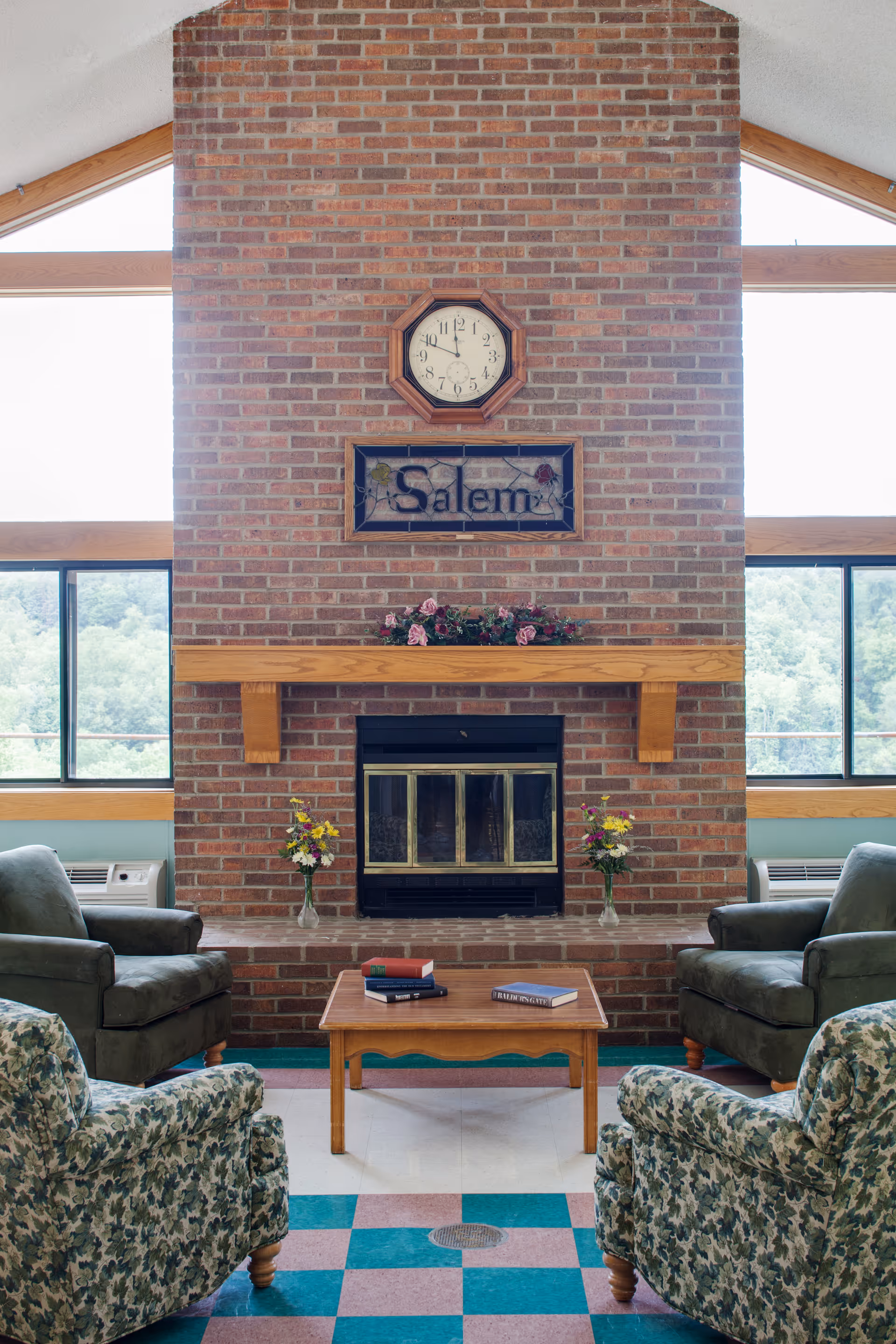 A sitting area with armchairs and patterned chairs arranged around a coffee table facing a brick fireplace with a clock and a framed 'Salem' sign above it.