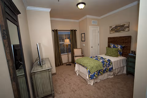 Cozy single bedroom with a twin bed, patterned bedding, dresser with a TV, floor lamp, and window with curtains.