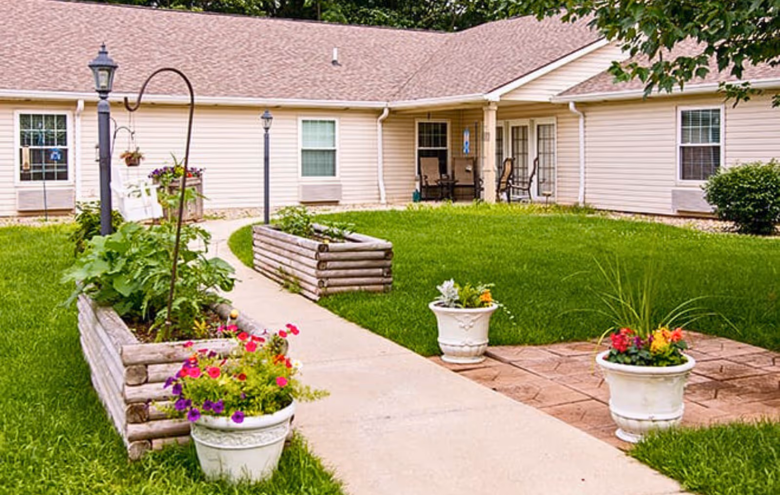 A well-maintained outdoor garden area with green grass, flower pots with colorful flowers, and raised wooden garden beds. A paved walkway leads to a building with beige siding and several windows. There are chairs and a small table on a covered porch area attached to the building.