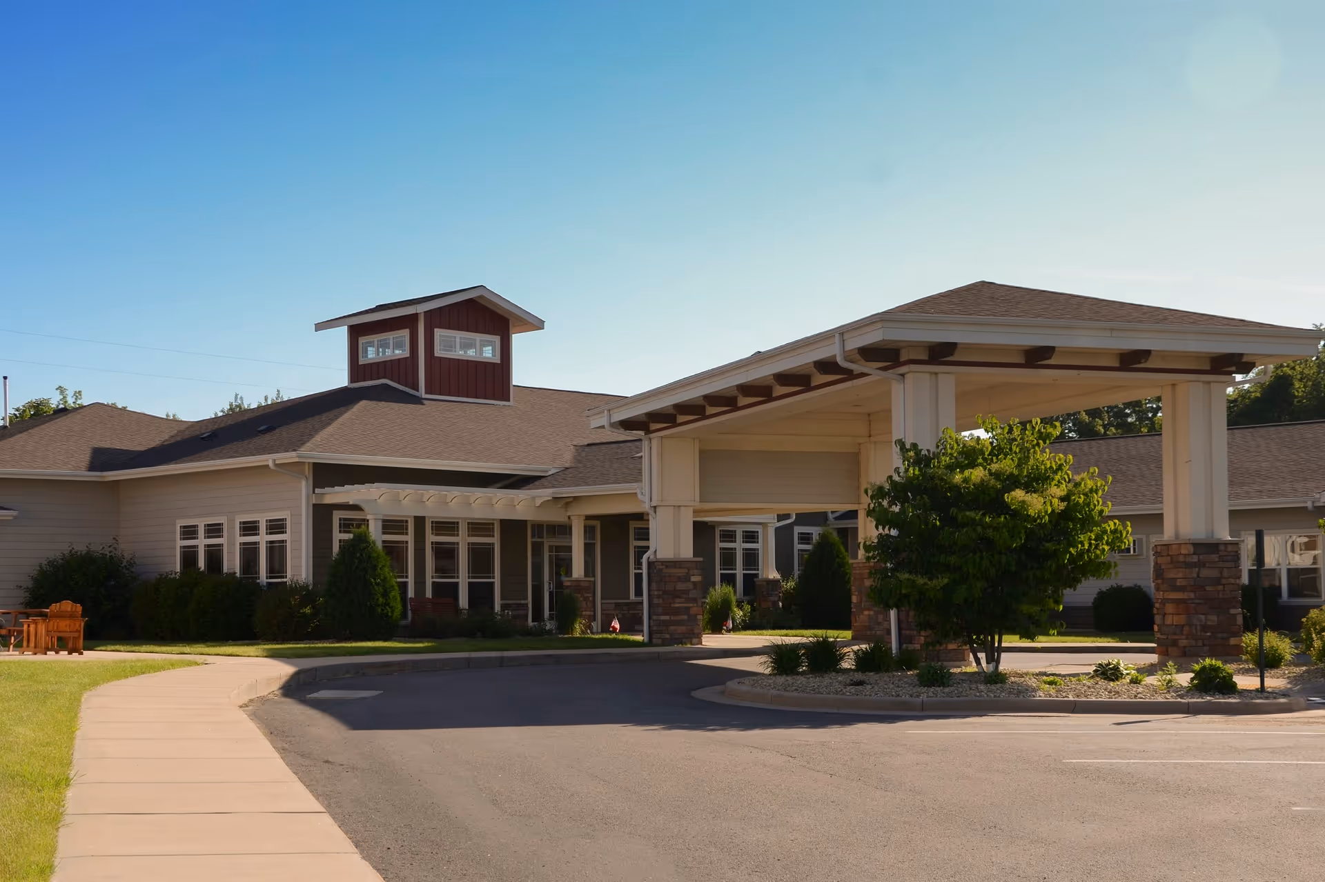 Exterior view of a senior living facility building with a covered entrance supported by white columns with brick bases. The building has a beige exterior with multiple windows and a small tower-like structure on the roof. There is a curved driveway and a sidewalk leading to the entrance, with some greenery and trees around the area under a clear blue sky.