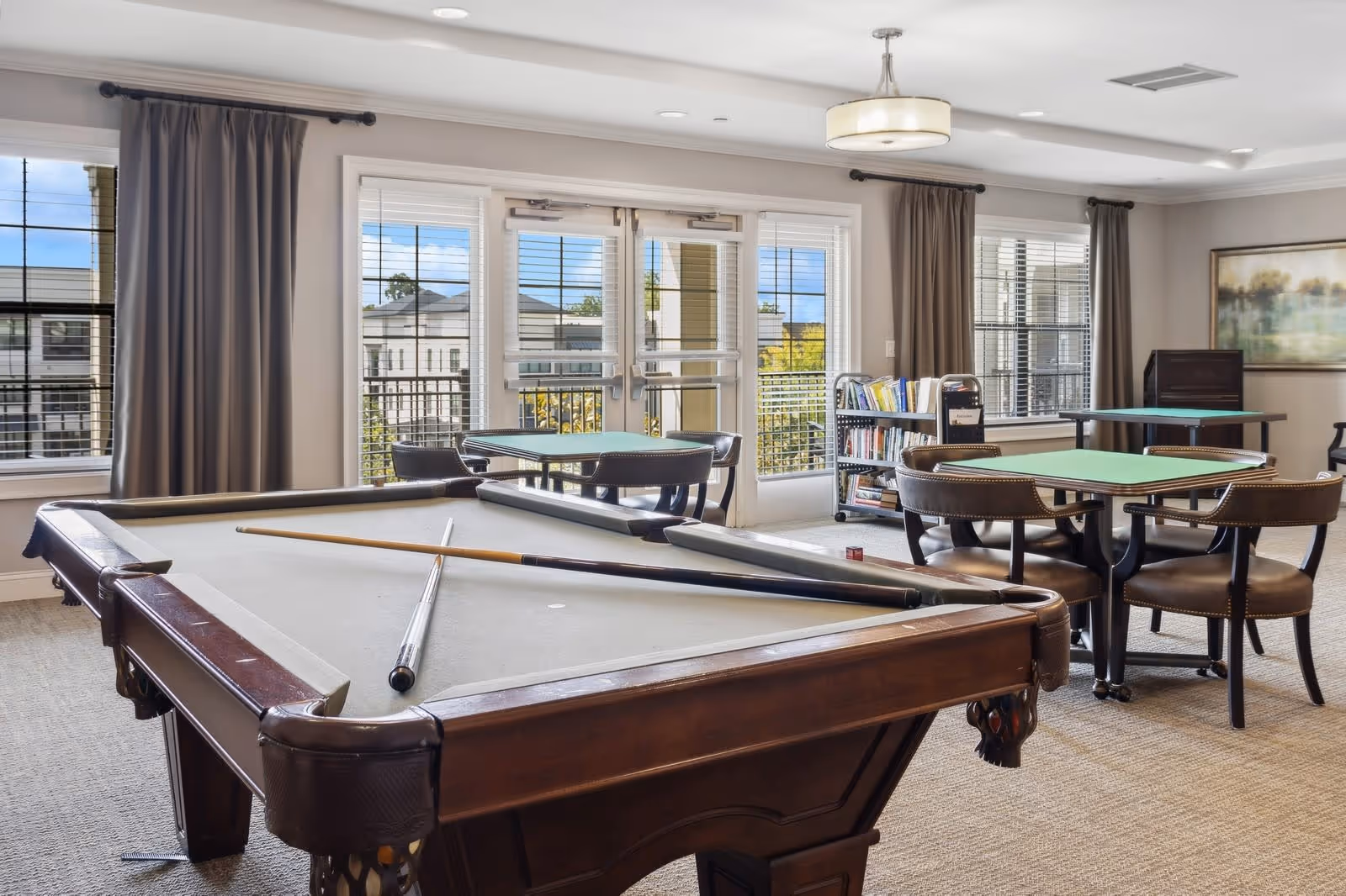 A bright game room with a pool table in the foreground, two pool cues resting on the table, and several card tables with chairs arranged around the room. Large windows and glass doors let in natural light, with curtains on either side. A bookshelf and framed artwork are visible in the background.