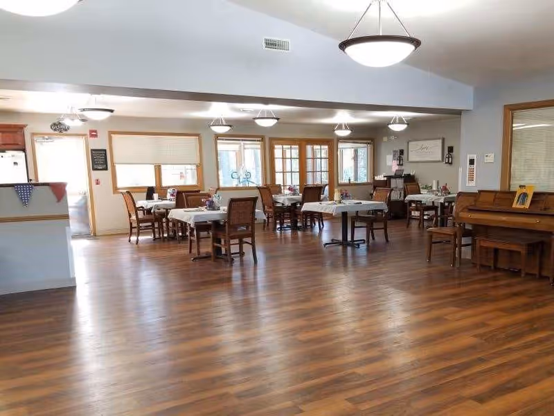 Spacious communal dining room with several set tables and chairs, wood floors, hanging lights, and a piano along the wall.