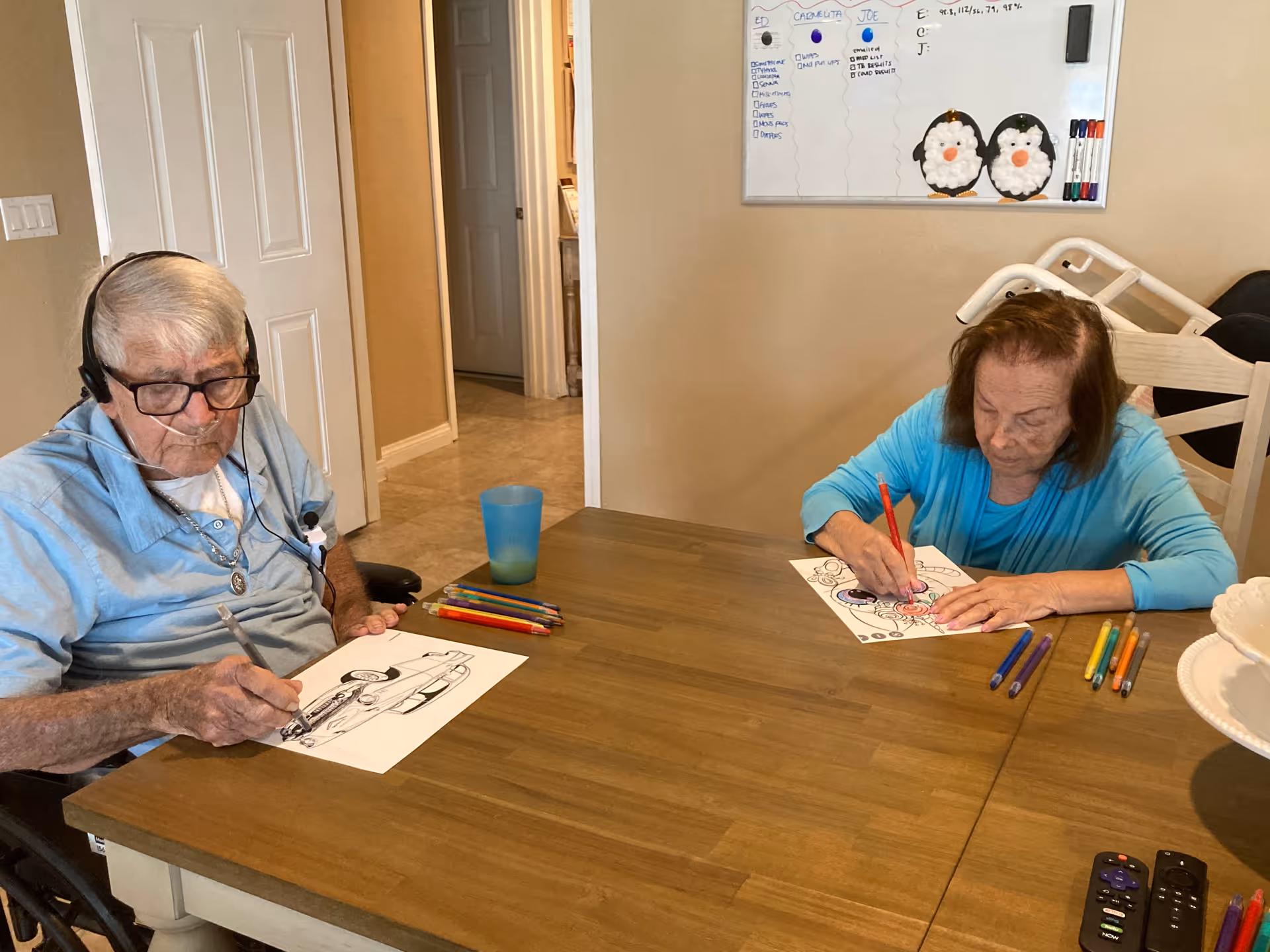 Two elderly individuals sitting at a wooden table in a room, engaged in coloring activities with colored pencils. One person is wearing headphones and glasses, coloring a picture of a car, while the other is focused on coloring a different picture. A whiteboard with notes and two penguin decorations is visible on the wall behind them.