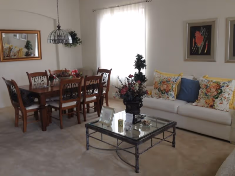 A cozy living and dining area with a wooden dining table and six chairs on the left, a glass coffee table with a floral arrangement in the center, and a white sofa with colorful floral and blue cushions on the right. The room is softly lit by natural light coming through a window with sheer white curtains. There are framed artworks on the wall above the sofa and a mirror on the wall near the dining table.