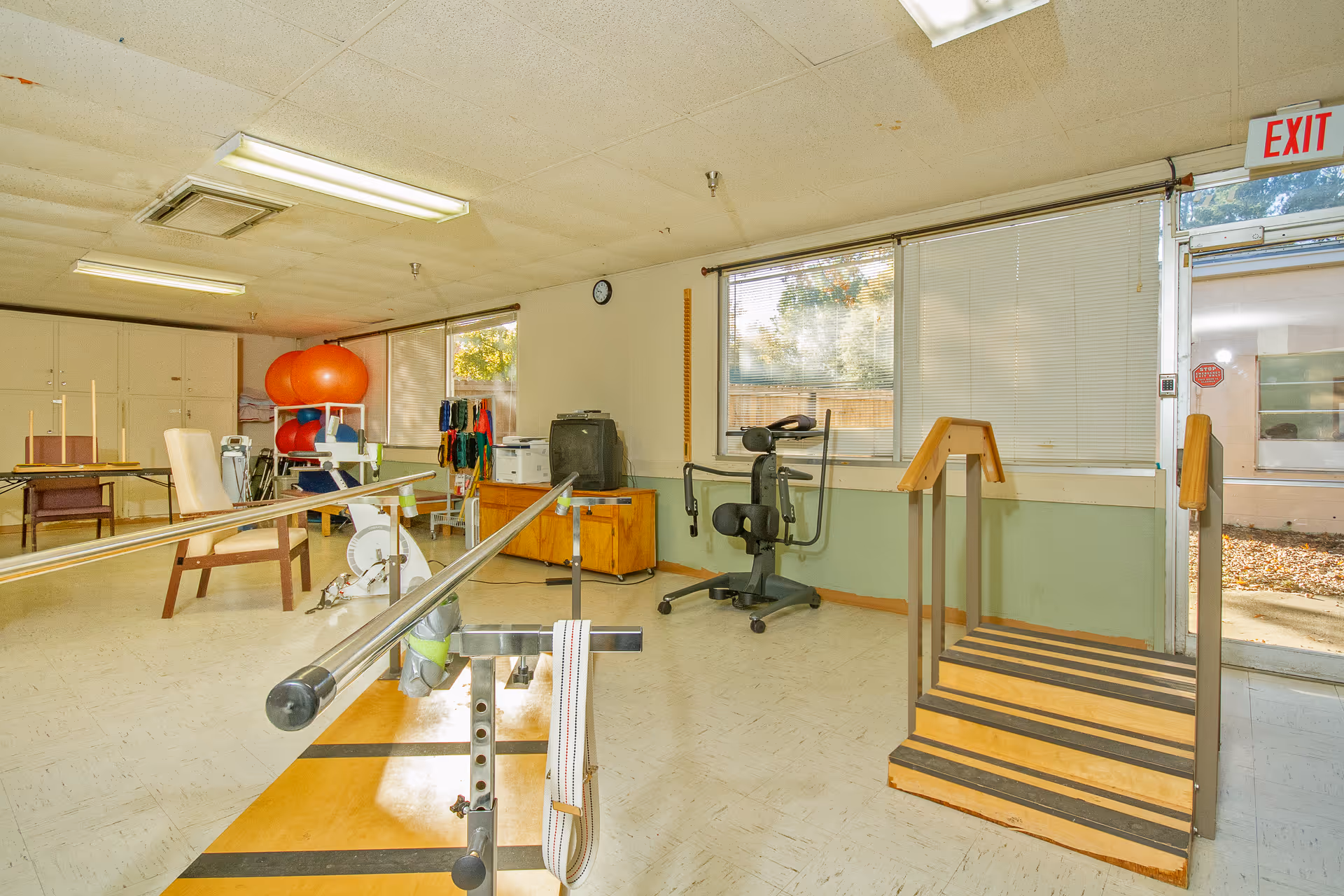 A rehabilitation room with parallel bars for walking exercises, a small set of wooden stairs with handrails, exercise equipment including an exercise bike, therapy balls, and resistance bands. The room has large windows with blinds, a clock on the wall, and an exit door leading outside.