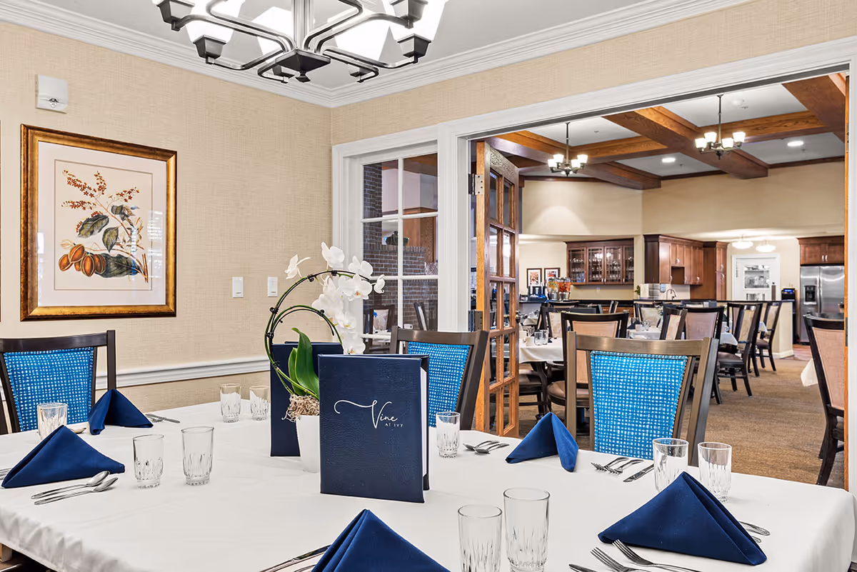 A dining room with a table set for a meal, featuring blue napkins, clear drinking glasses, silverware, and a centerpiece with white orchids. The room has beige walls, a framed botanical print, and blue cushioned chairs. In the background, another dining area with wooden beams on the ceiling and more tables and chairs is visible.