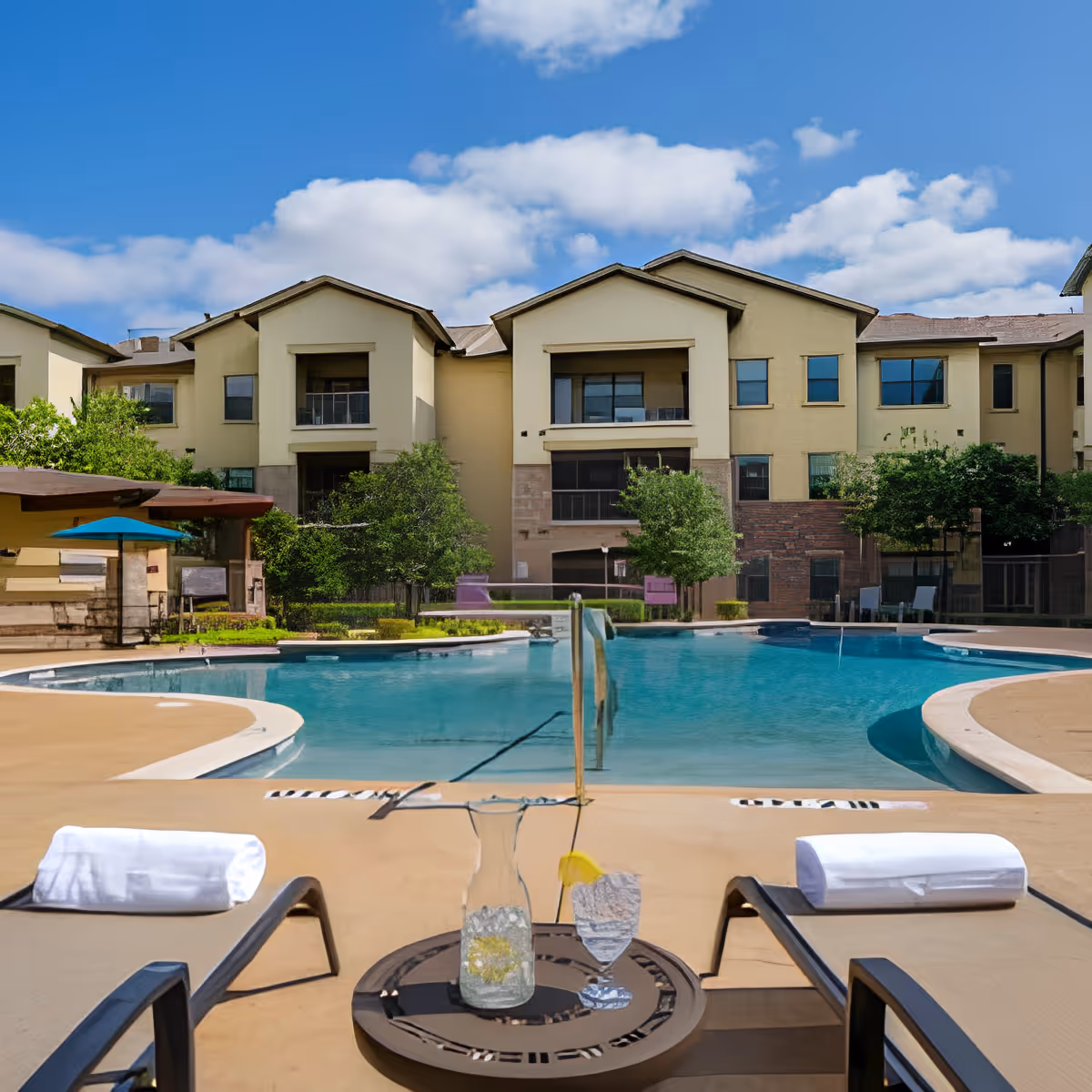 Outdoor swimming pool area with lounge chairs and a small table holding a pitcher of water and a glass with a lemon slice. In the background, there is a multi-story residential building with balconies and windows, surrounded by trees and greenery under a blue sky with scattered clouds.