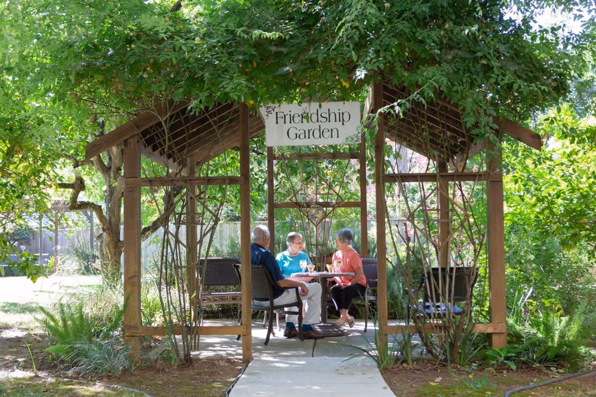 Three elderly people sitting and chatting at a table under a wooden gazebo labeled 'Friendship Garden' surrounded by lush green plants and trees.