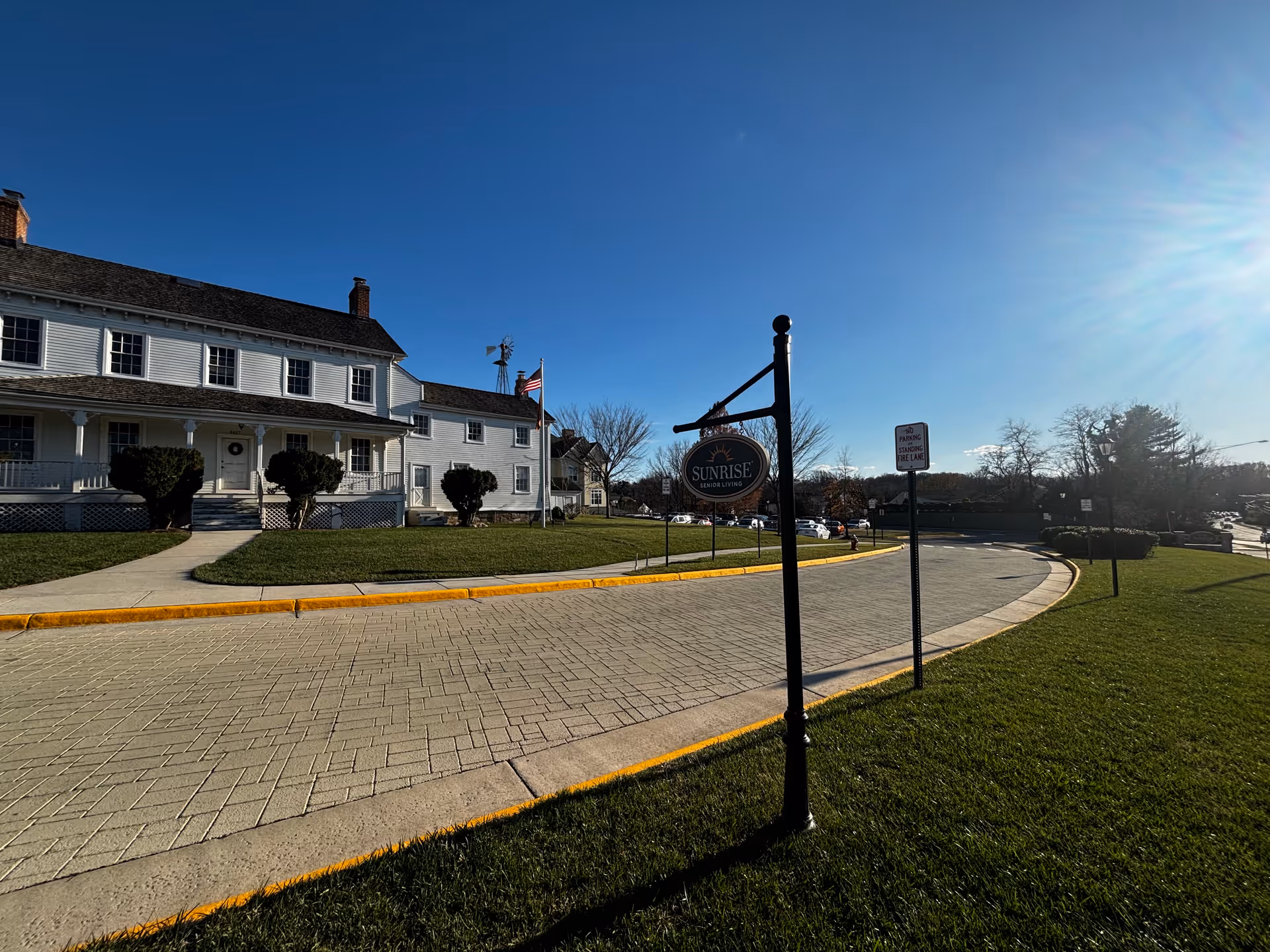 Exterior view of Sunrise at Silas Burke House senior living facility on a sunny day, showing a large white building with multiple windows, a porch, and a paved driveway with a signpost displaying the Sunrise Senior Living logo. There is a well-maintained lawn and clear blue sky.