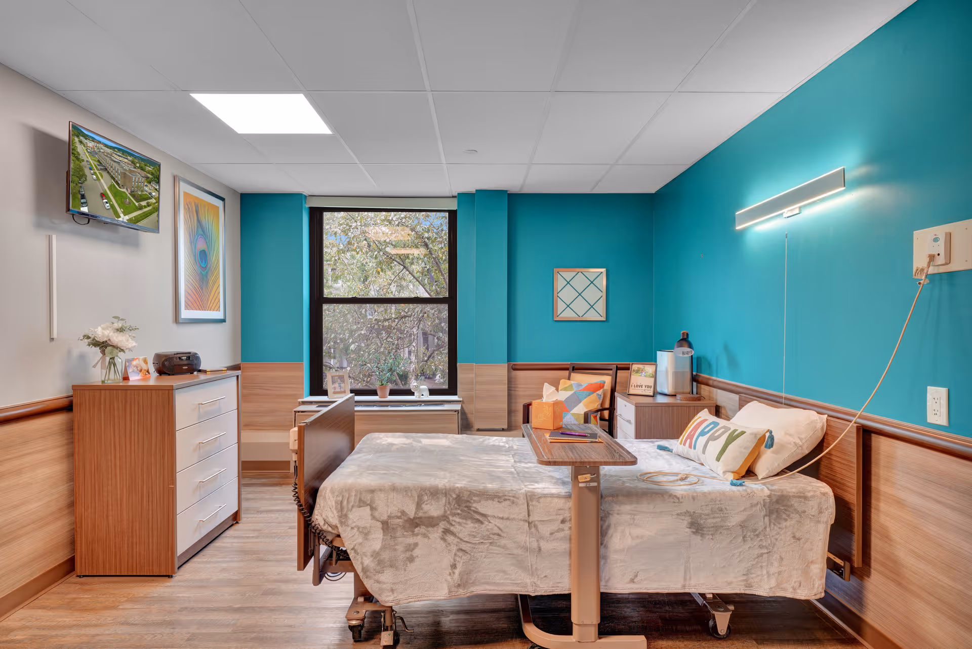A bright and cozy patient room in a rehabilitation and nursing facility featuring a hospital bed with a beige blanket and a pillow with the word 'HAPPY' on it. The room has teal walls with wood paneling, a large window showing trees outside, a wooden dresser with a vase of flowers, a mounted TV, and a small table on wheels positioned over the bed. There are colorful framed artworks on the walls and a chair with a colorful cushion next to a small cabinet.