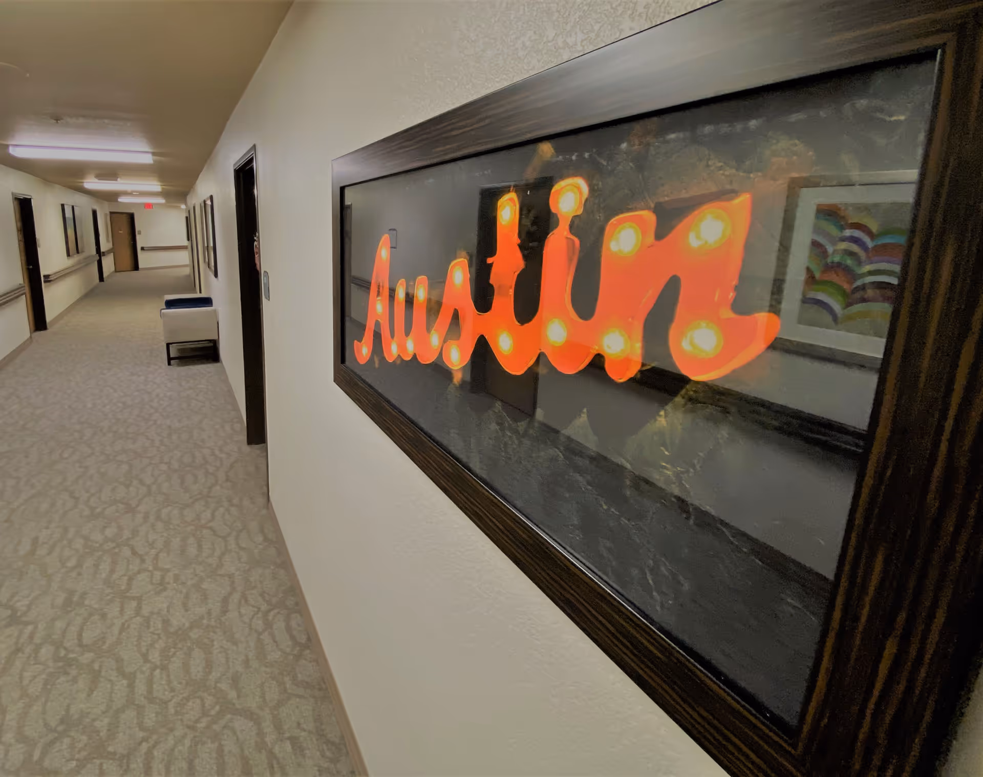 A long indoor hallway in a senior living facility with beige carpet and white walls. On the right wall, there is a framed artwork with the word 'Austin' in bright orange lights. The hallway has several doors and a bench along the left side, with ceiling lights illuminating the space.
