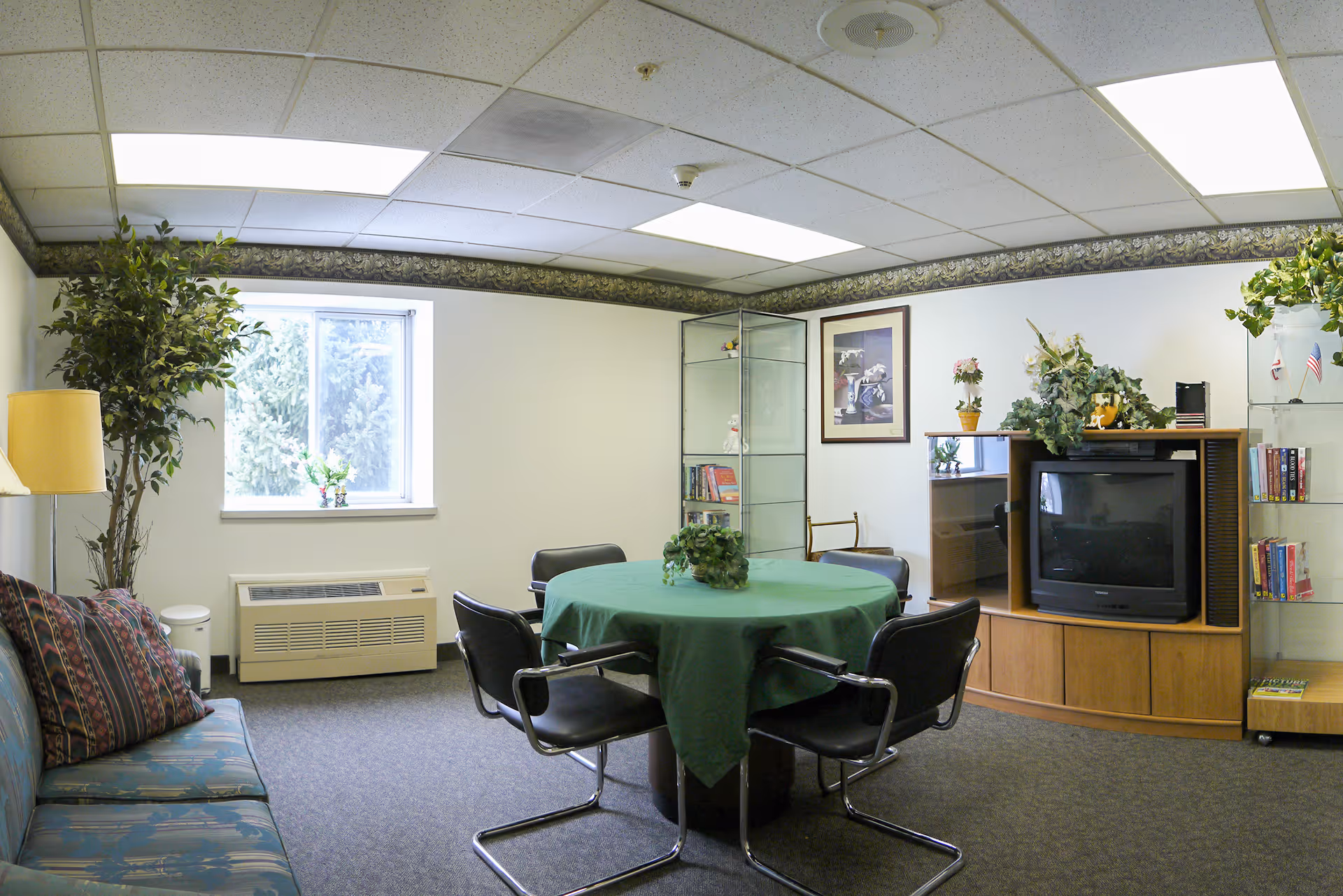 A cozy common room with a round table covered by a green tablecloth surrounded by four black chairs. There is a patterned couch with pillows on the left side, a tall potted plant near a window, and a wooden entertainment center with an old-style television and decorative plants. The room has carpeted flooring, a drop ceiling with fluorescent lights, and glass display cabinets with books and small decorations.
