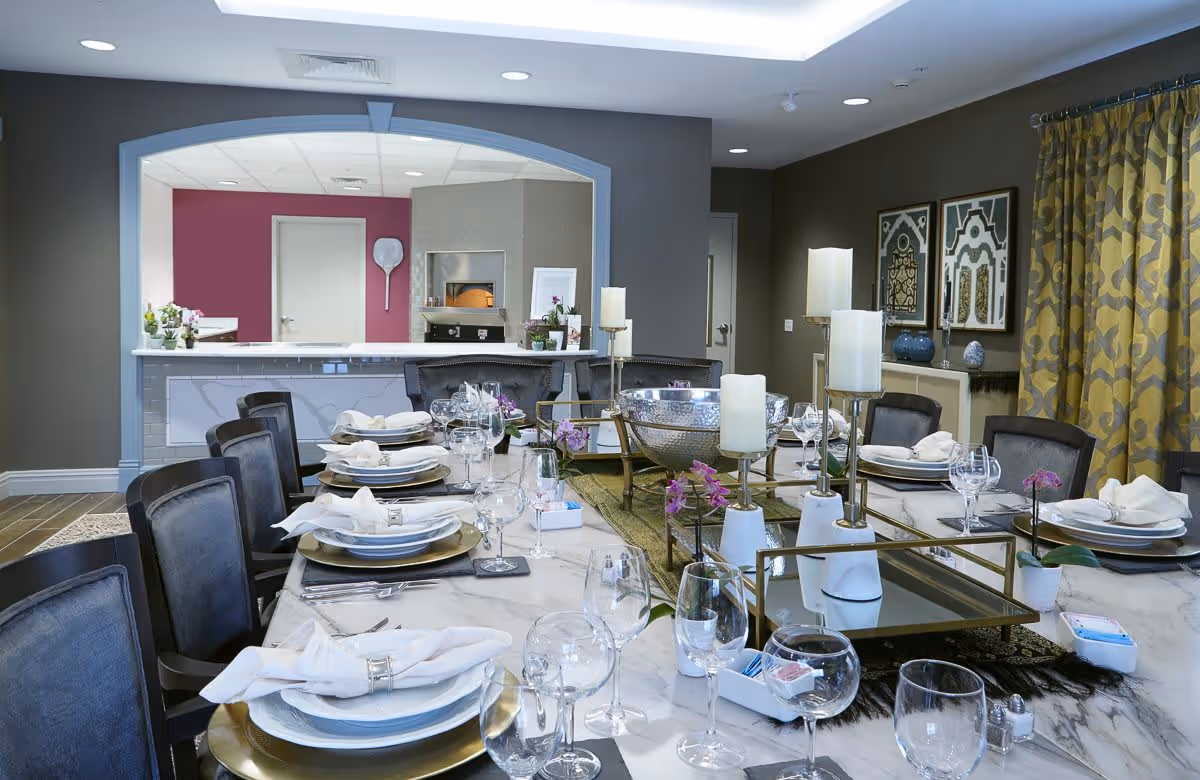 A formal dining room with a long marble table set for a meal, featuring white plates, folded napkins, wine glasses, and silverware. The table is decorated with candles, small purple flowers, and a large silver bowl centerpiece. The room has dark walls, patterned yellow curtains, framed artwork, and an open kitchen area in the background with a pizza oven.