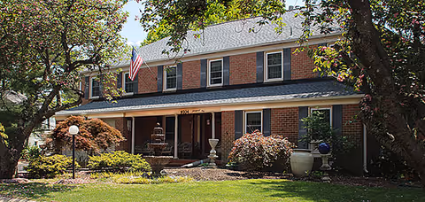 Front exterior view of a two-story brick assisted living facility with multiple windows, an American flag, a central entrance with a covered porch, a water fountain in the front yard, and landscaped greenery including trees and bushes.