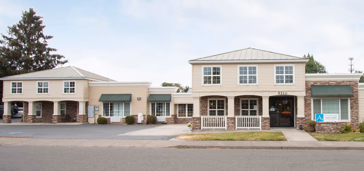 Exterior front view of Avamere Court at Keizer, a two-story beige building with stone accents, multiple windows, green awnings, and a main entrance with the address number 5210.