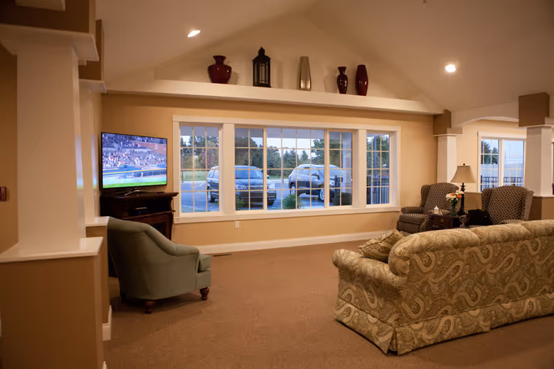 A warm assisted-living common room with a patterned sofa and armchairs facing a TV and a large multi-pane window overlooking parked cars.