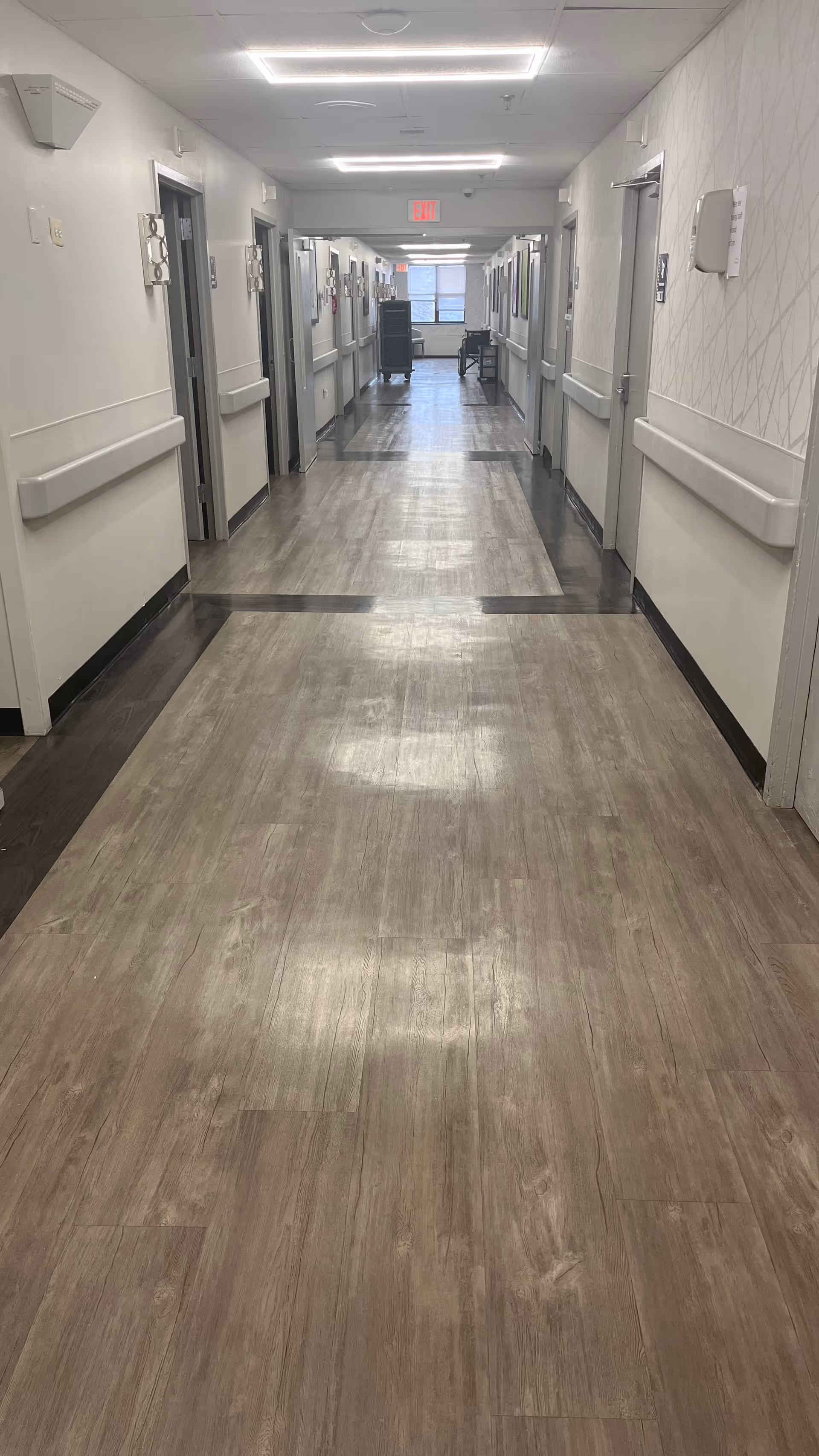 Long interior corridor with wood-look flooring, handrails and doors to rooms on both sides under ceiling lights.
