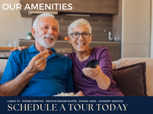 An elderly man and woman sitting on a couch in a living room, smiling and enjoying watching TV together. The woman is holding a remote control and the man is holding popcorn. The background shows a kitchen area.