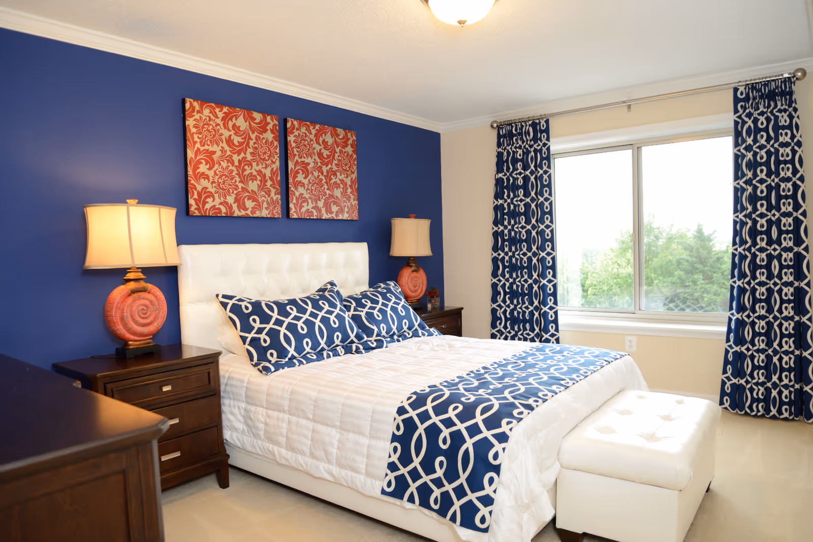 A bright bedroom with a white tufted bed featuring blue and white patterned pillows and a matching bed runner. The wall behind the bed is painted blue and decorated with two red and orange patterned artworks. There are two wooden nightstands with matching lamps that have orange spiral bases. A large window with blue and white patterned curtains lets in natural light, and a white upholstered bench sits at the foot of the bed.