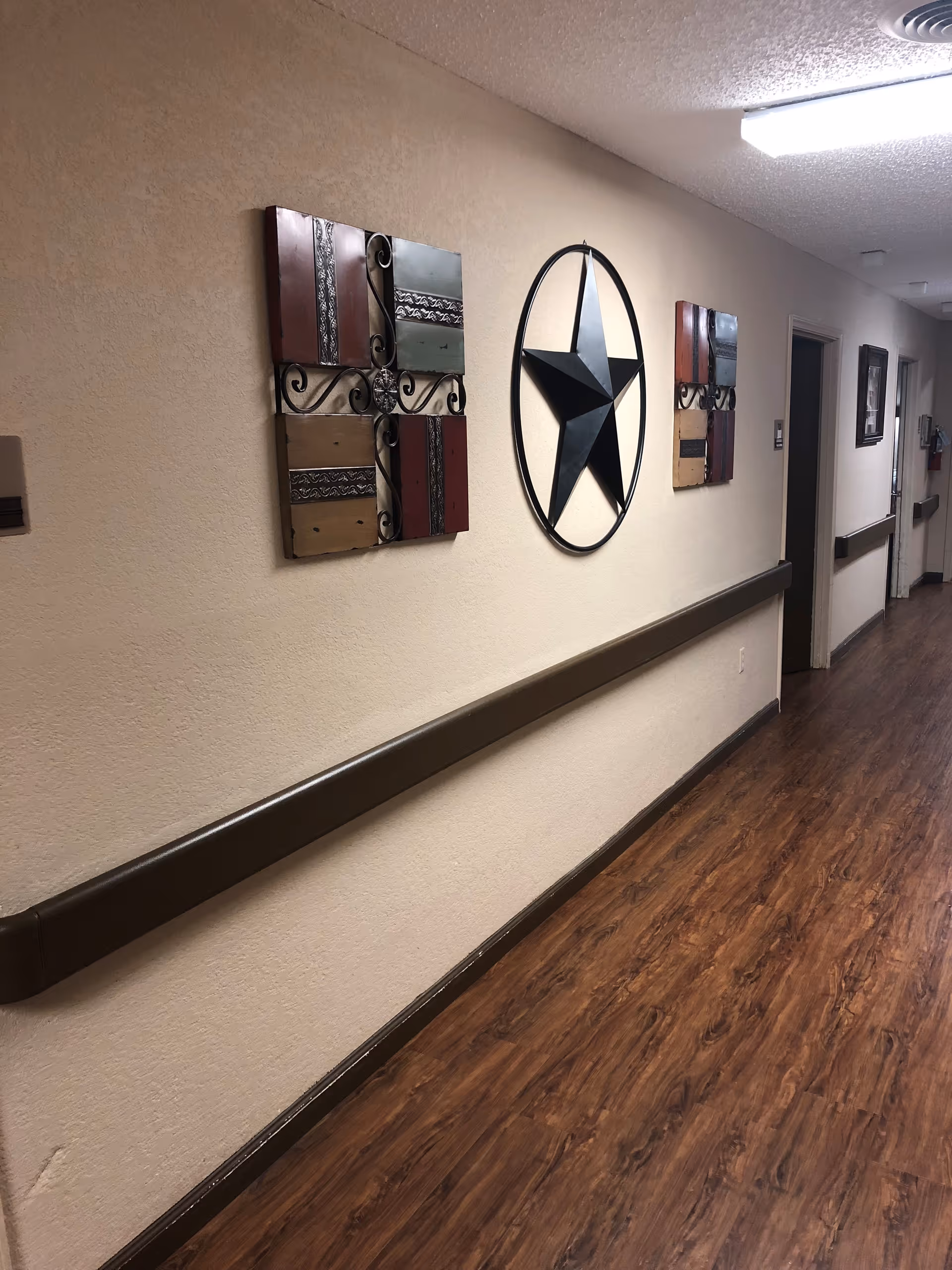 A hallway with wood-patterned flooring and beige walls featuring decorative wall art including a large black star and colorful square panels. The hallway has handrails along the walls and several doors leading to rooms.