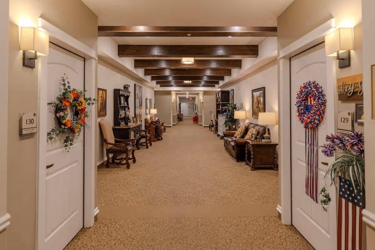 A long, well-lit hallway in an assisted living facility with beige walls and carpet. The hallway features wooden ceiling beams, wall sconces, and doors decorated with wreaths. Along the walls are chairs, tables with lamps, framed artwork, and plants, creating a warm and inviting atmosphere.