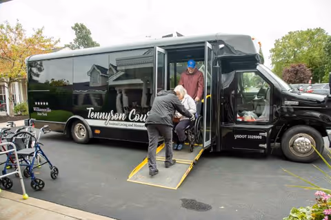A black shuttle bus with the name Tennyson Court on the side is parked on a driveway. A person is assisting an elderly woman in a wheelchair as she is being helped off the bus via a ramp. Another person stands inside the bus. Several walkers are visible on the left side of the image, and trees and houses are in the background.