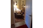 Partially open door leading into a cozy sitting area with an upholstered armchair, floor lamp, bookshelf, framed pictures, and an area rug.