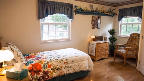 A cozy bedroom with a single bed covered in a floral bedspread, a wooden nightstand with a lamp and tissue box, two windows with blue curtains, a wooden dresser with decorative items, a framed photo collage on the wall, a potted plant, and a cushioned armchair with a floral pattern.