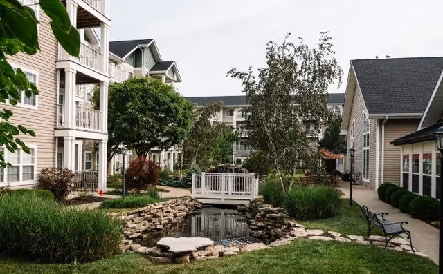 Outdoor courtyard area at a senior living facility featuring a small pond with a white wooden bridge over it, surrounded by landscaped greenery, trees, benches, and multi-story residential buildings.