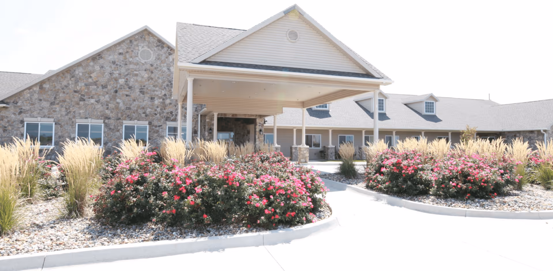 Front exterior view of Homestead Assisted Living & Memory Care of Osceola featuring a stone and siding building with a covered entrance, surrounded by landscaped bushes with pink flowers and ornamental grasses.