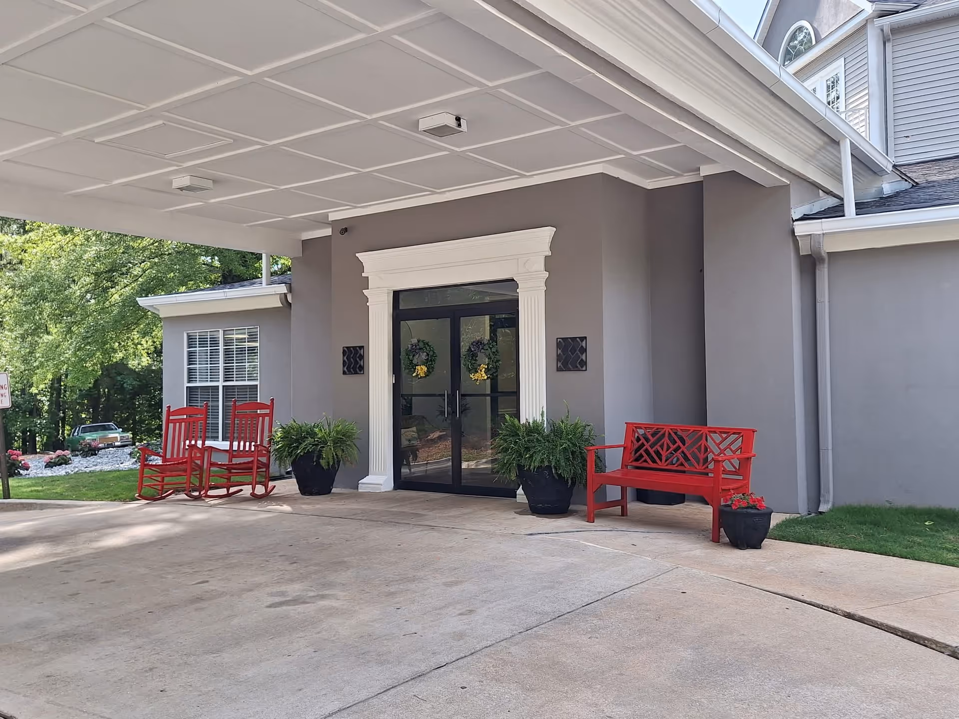 Entrance of a building with a covered driveway, featuring two red rocking chairs and a red bench on either side of double glass doors decorated with wreaths. There are potted plants near the doors and greenery in the background.