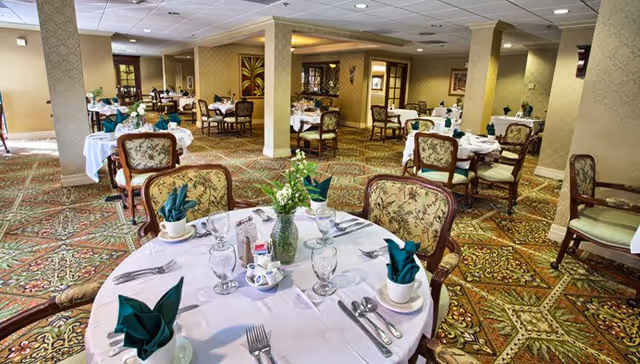 A spacious dining room with multiple round tables covered with white tablecloths, each set with glassware, silverware, cups, and green folded napkins. The room features patterned carpet, upholstered chairs with floral designs, and soft lighting. There are decorative elements on the walls and a vase with flowers on the table in the foreground.