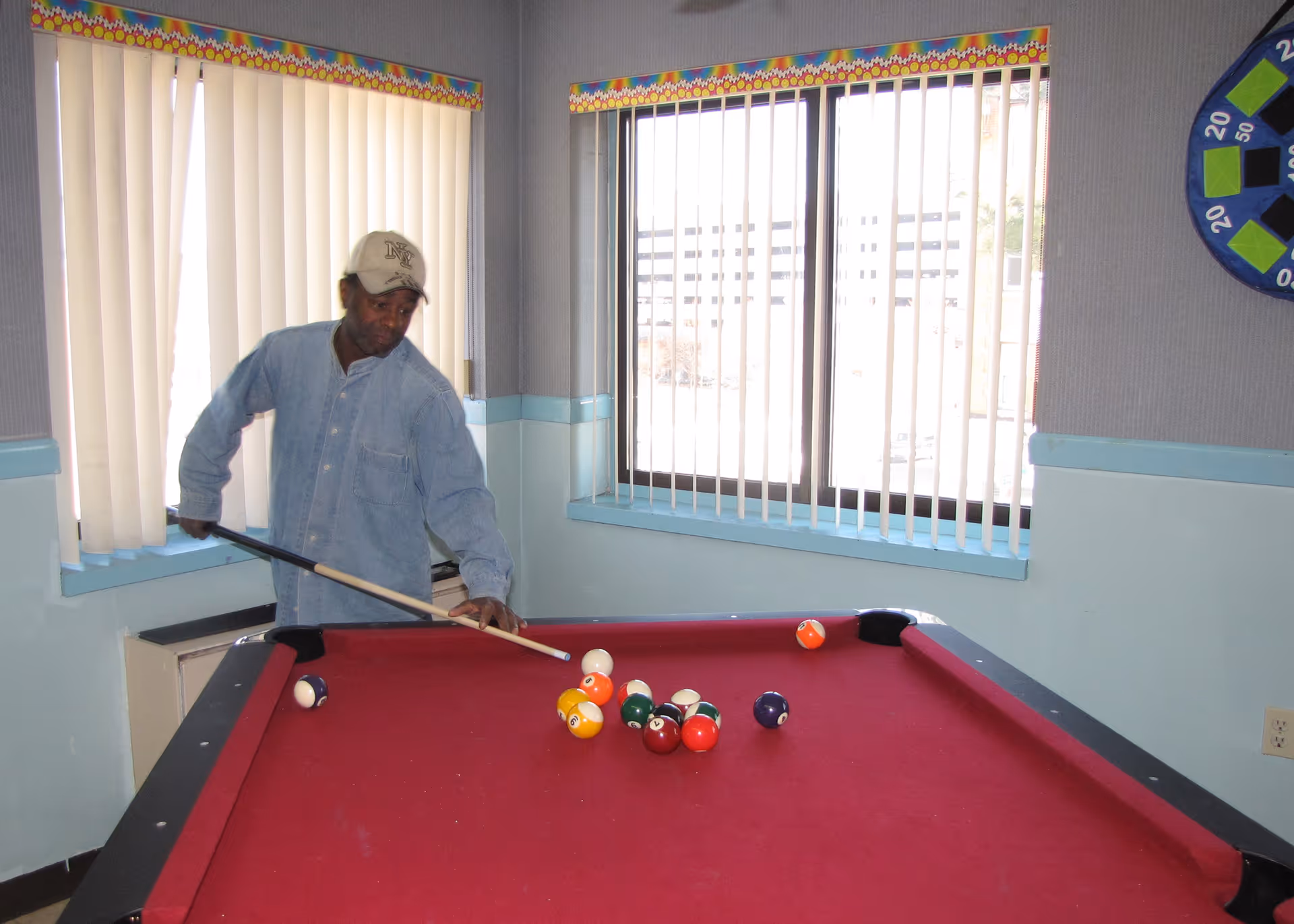 A man wearing a light blue shirt and a beige cap is playing pool on a red pool table in a room with light blue walls and vertical blinds on the windows.