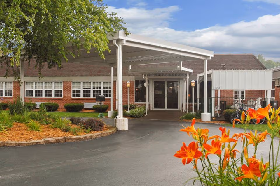 Entrance of a single-story brick building with a covered driveway, landscaped garden with orange flowers in the foreground, and a partly cloudy sky.