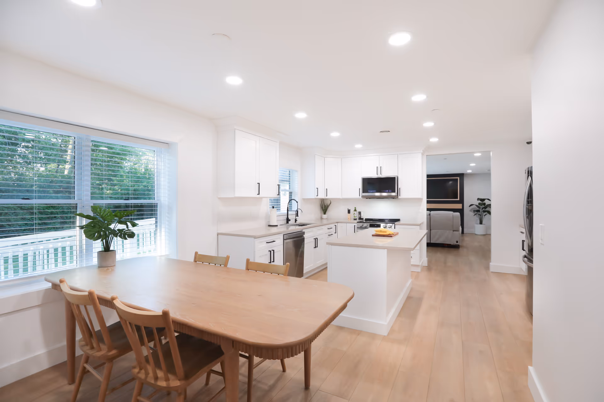 Bright and modern kitchen and dining area with white cabinetry, stainless steel appliances, a kitchen island, wooden dining table with four chairs, and large windows with blinds letting in natural light.