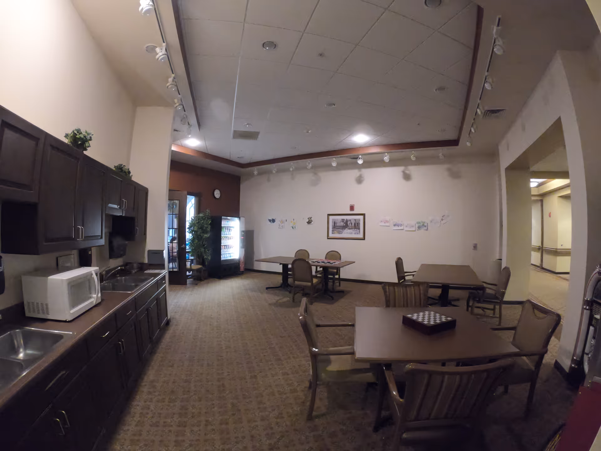 A senior living common room with a kitchenette, microwave, vending machine, and several tables and chairs arranged for dining or activities.