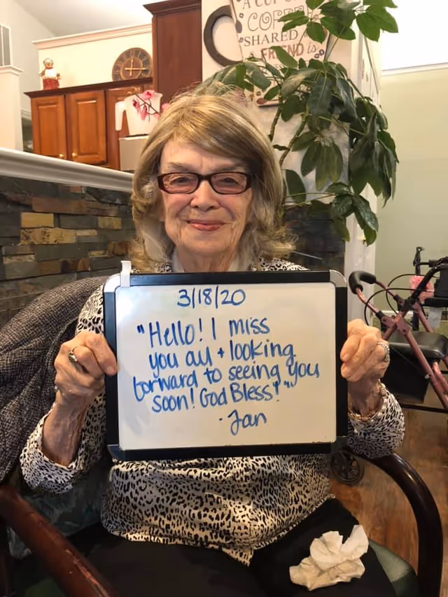 An elderly woman with glasses and light brown hair is sitting in a chair indoors, holding a whiteboard with a handwritten message dated 3/18/20 that says, 'Hello!! I miss you all + looking forward to seeing you soon! God Bless! - Jan'. Behind her is a stone wall, wooden cabinets, a large green plant, and a pink walker.