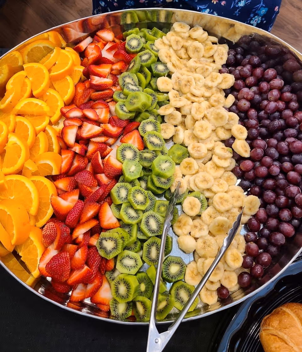 A large round tray filled with neatly arranged fresh fruit slices including orange wedges, strawberries, kiwi slices, banana slices, and red grapes, with a pair of metal tongs resting on the tray.