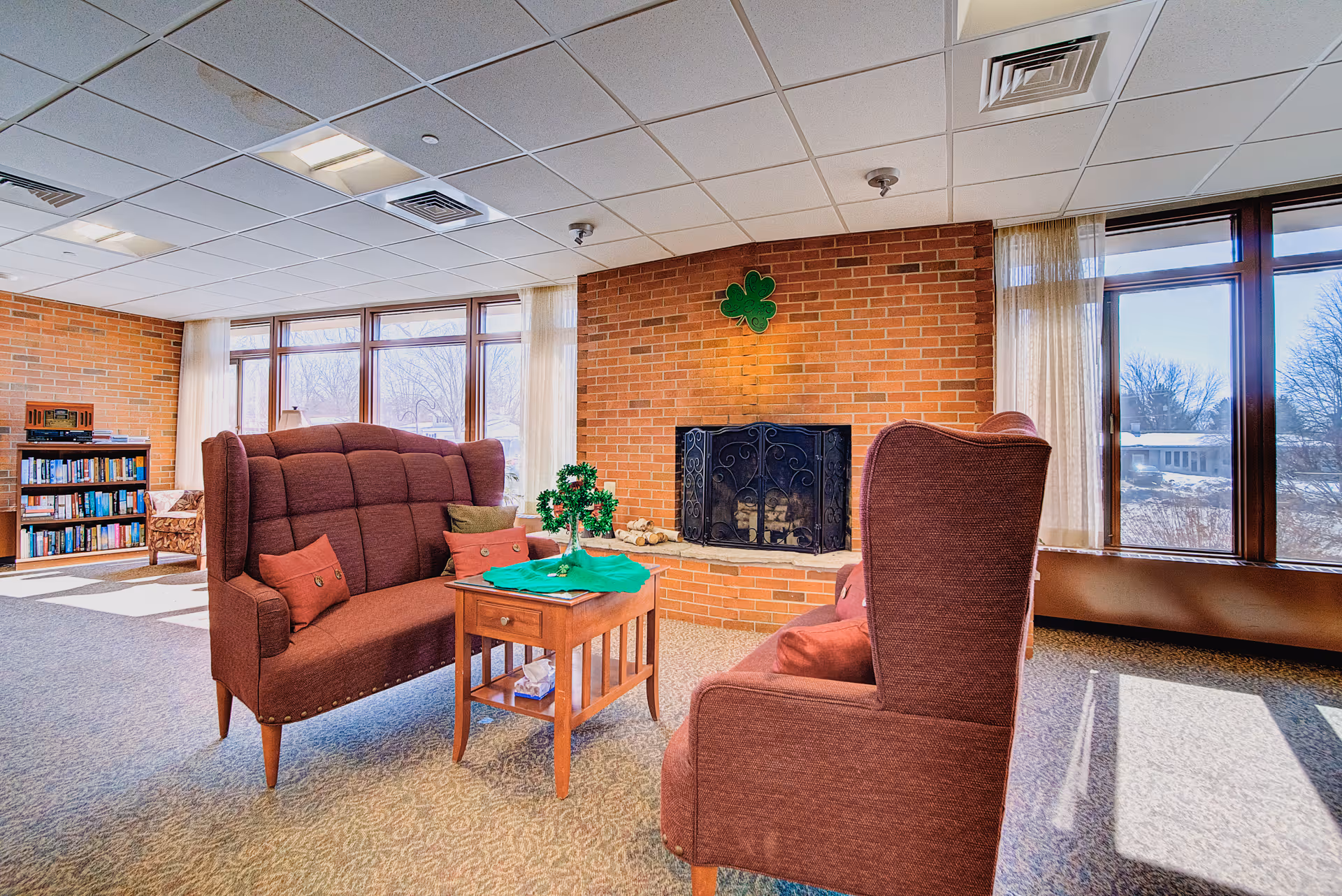 A cozy common area with two maroon upholstered armchairs and a matching loveseat arranged around a small wooden table. The table has a green shamrock decoration on it. Behind the seating is a brick fireplace with a decorative black metal screen and a green shamrock hanging above it. Large windows with sheer curtains let in natural light, and a bookshelf filled with books is visible in the background.