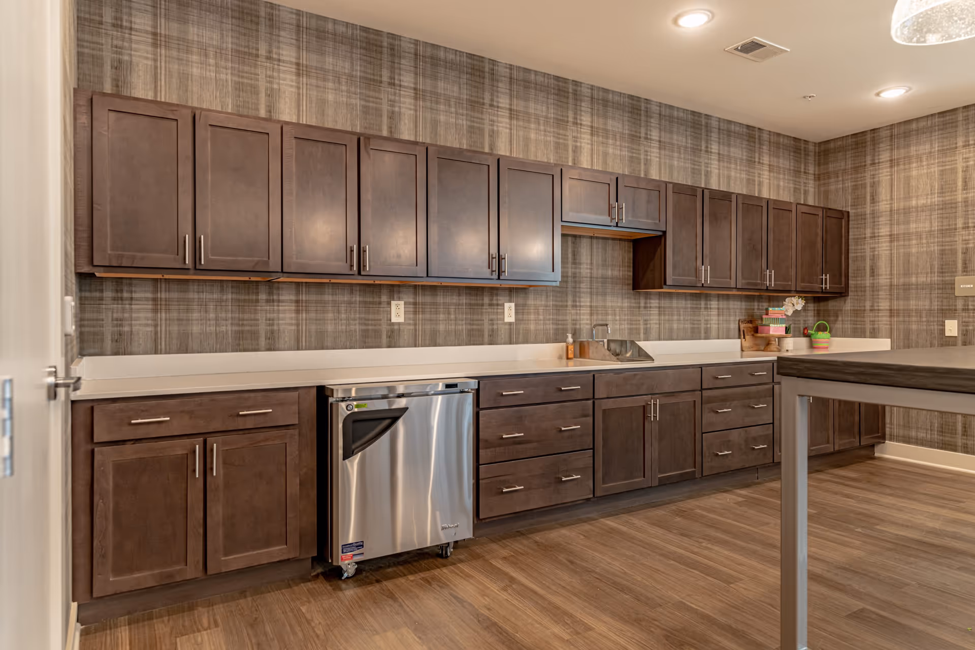 A modern kitchen with dark wooden cabinets, a stainless steel dishwasher, a small sink, and a countertop with a few decorative items. The walls have a textured, plaid-patterned wallpaper, and the floor is a wood finish. The ceiling has recessed lighting.