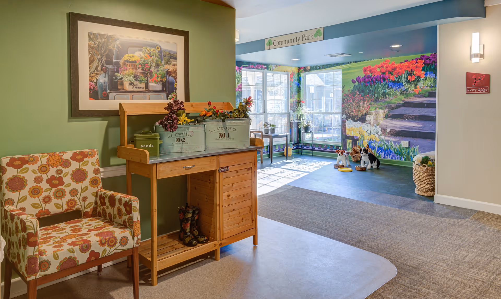 A bright and welcoming indoor community space with a floral patterned armchair next to a wooden storage cabinet holding decorative flower pots and gardening boots. The walls are painted green and beige, with a large framed picture of flowers above the cabinet. In the background, there is a colorful mural of a garden with flowers and stone steps, large windows letting in natural light, and a small play area with dog statues and bowls. A sign above the windows reads 'Community Park'.