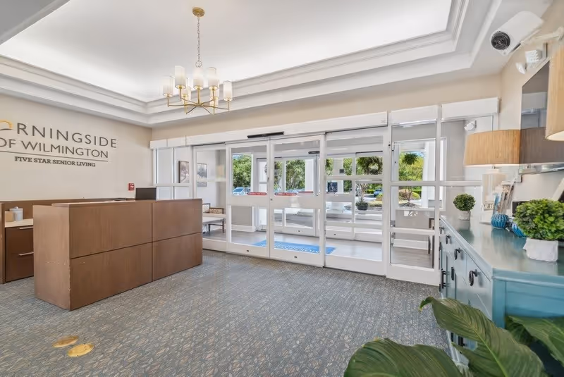 Reception lobby with a wooden front desk, automatic glass entrance doors, plants, and a 'Morningside of Wilmington' sign on the wall.