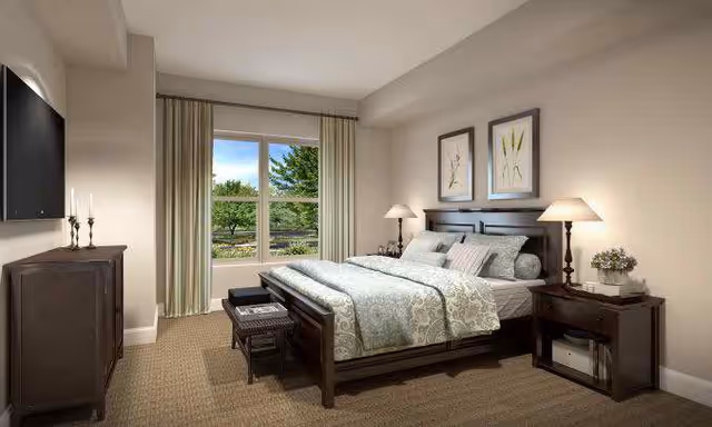 Bright, tastefully furnished bedroom with a wooden bed, matching nightstands and lamps, a dresser with wall-mounted TV, and a window showing greenery.