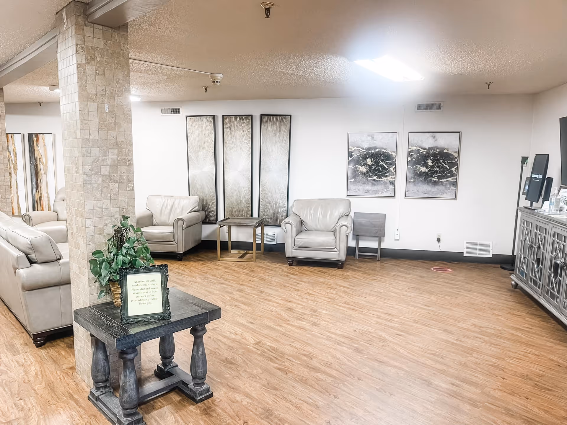 Spacious nursing home lounge with leather armchairs, side tables, wall art, and a small table with a potted plant in the foreground.