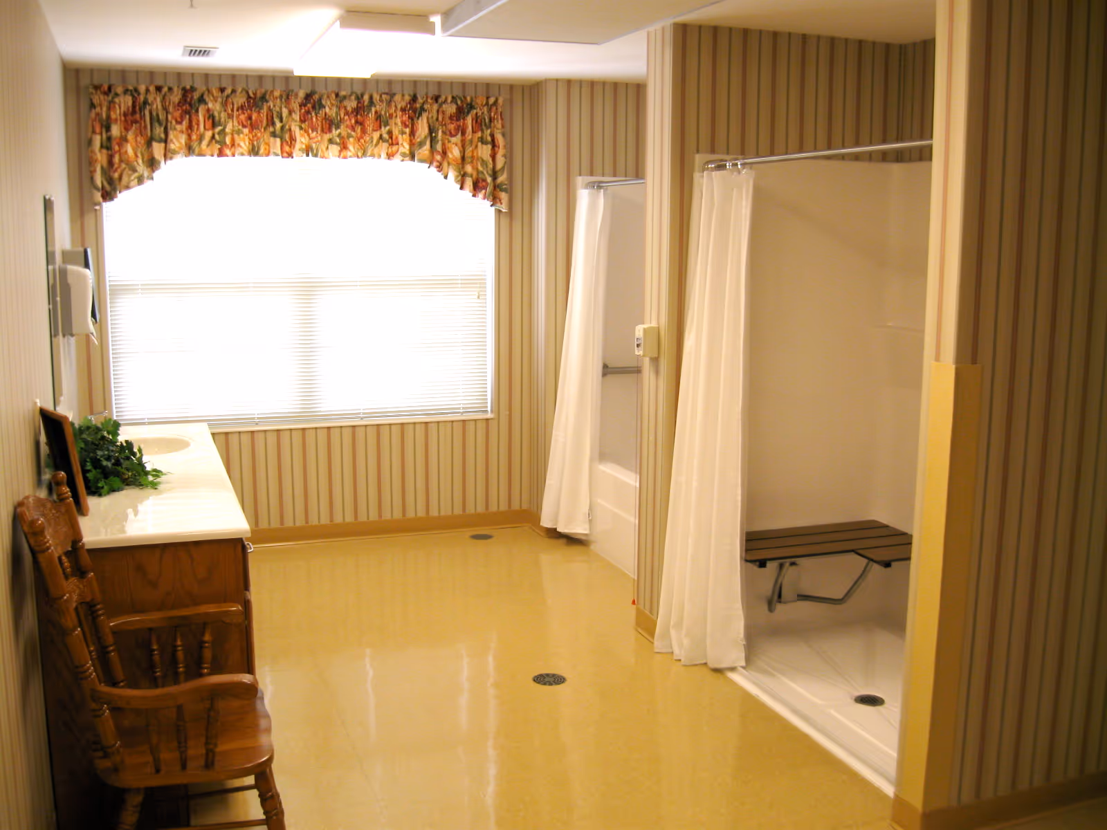 A spacious bathroom area with two shower stalls separated by a partial wall and white shower curtains. Each shower has a fold-down bench. There is a wooden chair and a long countertop with a sink on the left side. The room has striped wallpaper, a large window with floral valance, and a yellow linoleum floor.