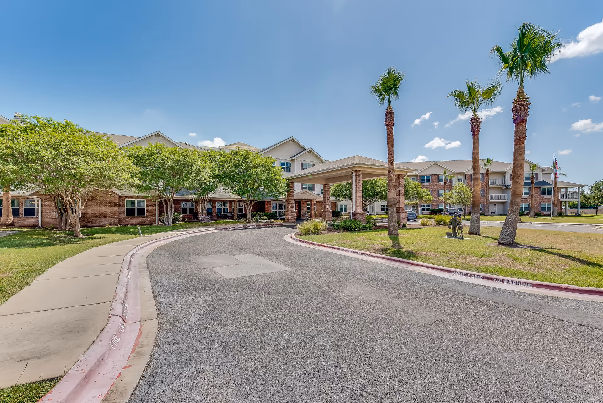Exterior view of Solstice Senior Living at Corpus Christi showing a curved driveway leading to the main entrance of a multi-story brick building with a covered drop-off area. There are palm trees and other greenery around the building under a clear blue sky.