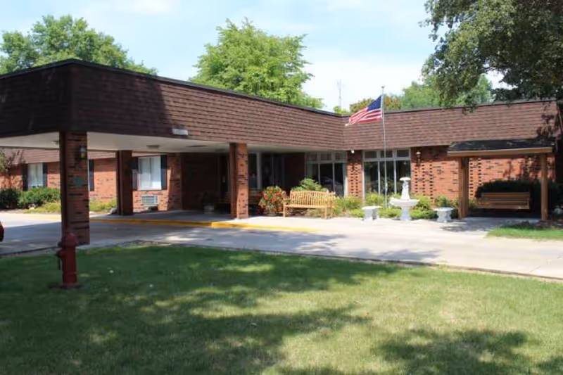 Exterior view of a single-story brick building with a covered entrance, benches, a small fountain, and an American flag on a flagpole. The area is surrounded by green grass and trees under a clear sky.
